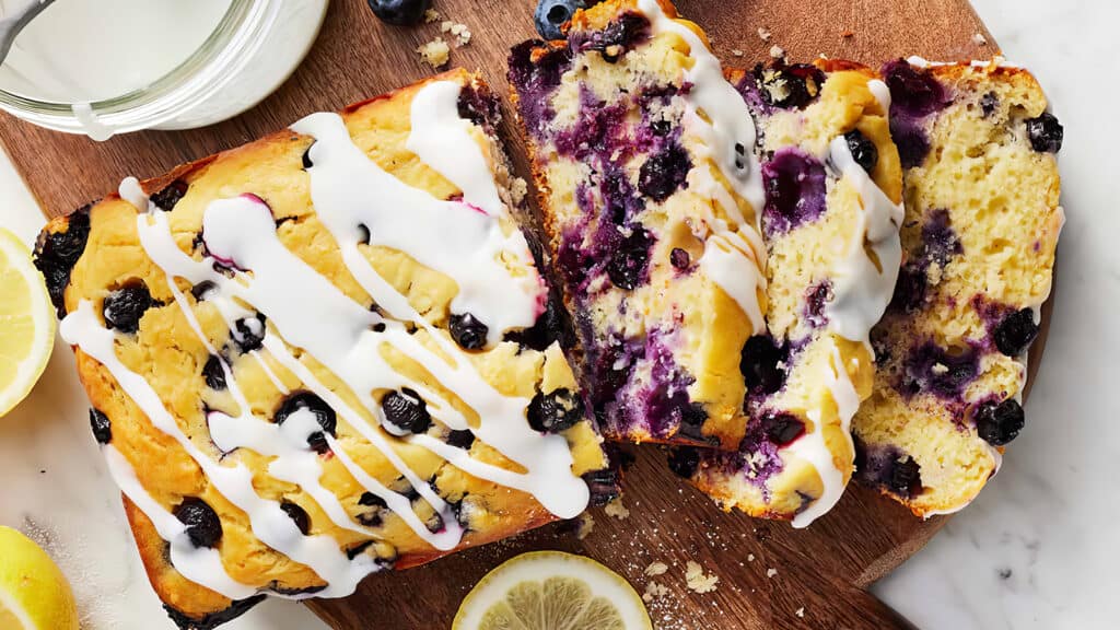 A loaf of blueberry lemon bread with white icing drizzled on top sits on a wooden cutting board. Two slices have been cut from the loaf, displaying blueberries and a moist texture inside. A lemon slice and a jar nearby add to the decorative presentation, perfect for exploring blueberry recipes.