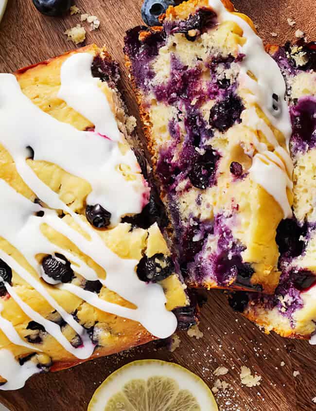 A loaf of blueberry lemon bread with white icing drizzled on top sits on a wooden cutting board. Two slices have been cut from the loaf, displaying blueberries and a moist texture inside. A lemon slice and a jar nearby add to the decorative presentation, perfect for exploring blueberry recipes.