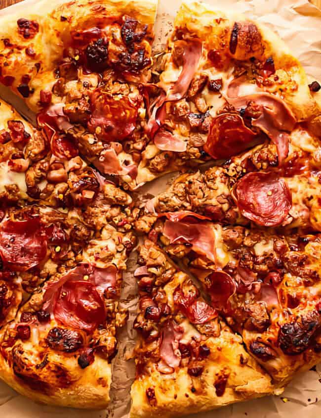 A delicious pizza with a crispy golden crust is topped with generous amounts of cheese, pepperoni, ham, and crumbled sausage. The pizza is sliced into eight pieces and placed on a sheet of parchment paper. Inspired by Asian recipes, a small bowl of chili flakes sits beside the pizza.