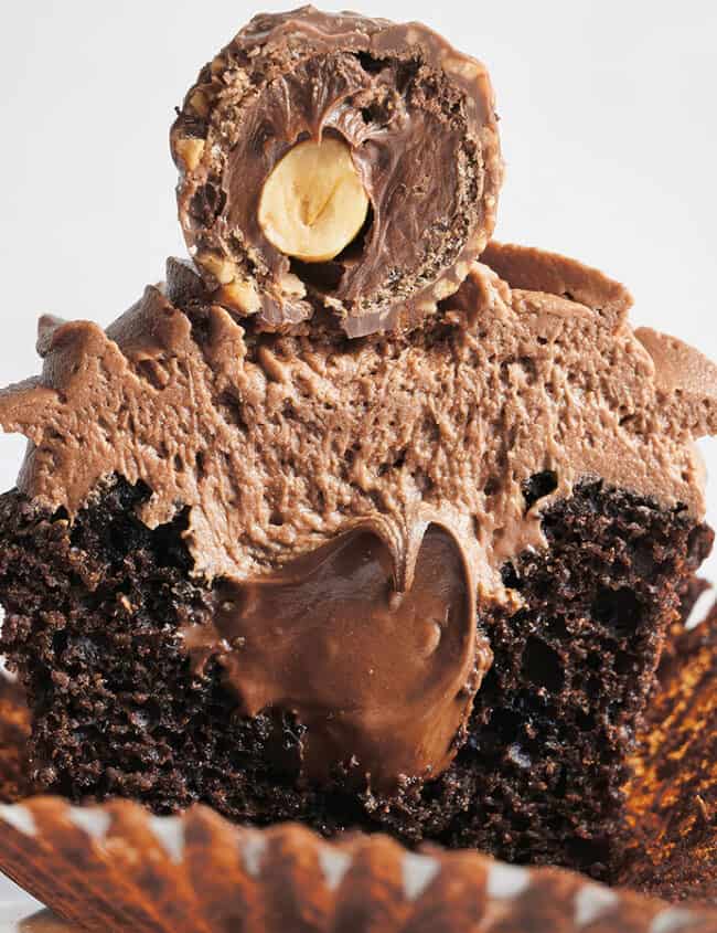 A decadent chocolate cupcake, one of the finest baked goods, topped with rich chocolate frosting and a Ferrero Rocher candy cut in half to reveal a creamy filling. The cupcake liner is peeled back, exposing the moist, dark chocolate cake. A delicious addition to any desserts table against a plain white background.