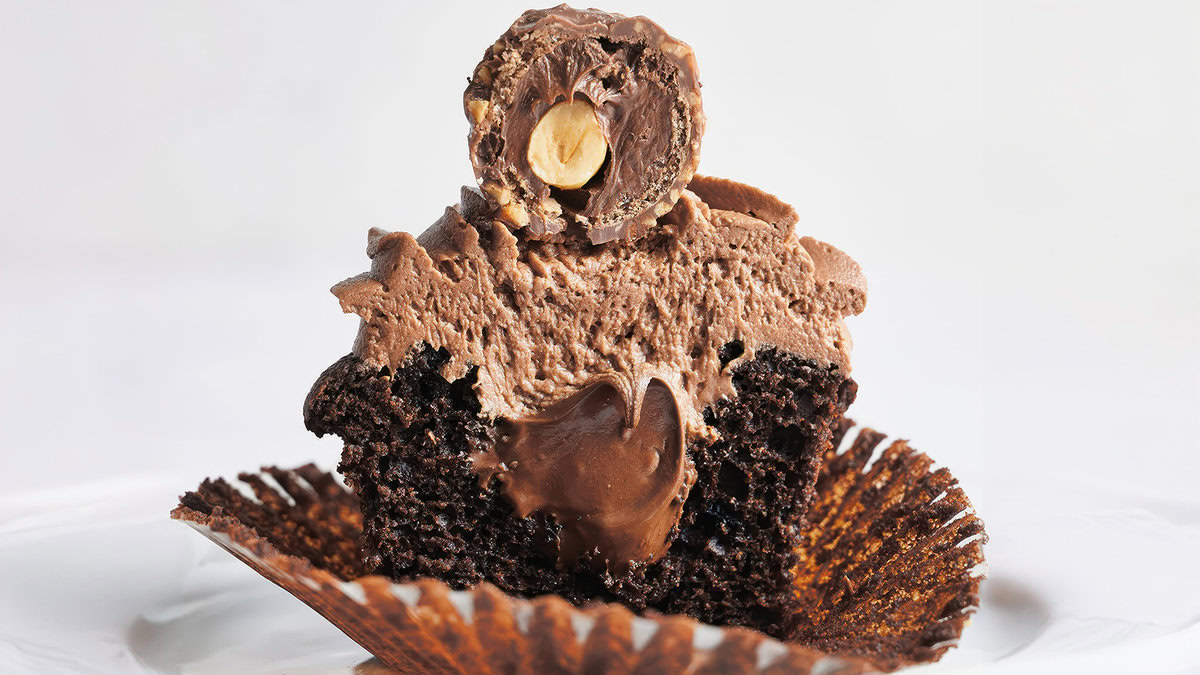 A decadent chocolate cupcake, one of the finest baked goods, topped with rich chocolate frosting and a Ferrero Rocher candy cut in half to reveal a creamy filling. The cupcake liner is peeled back, exposing the moist, dark chocolate cake. A delicious addition to any desserts table against a plain white background.