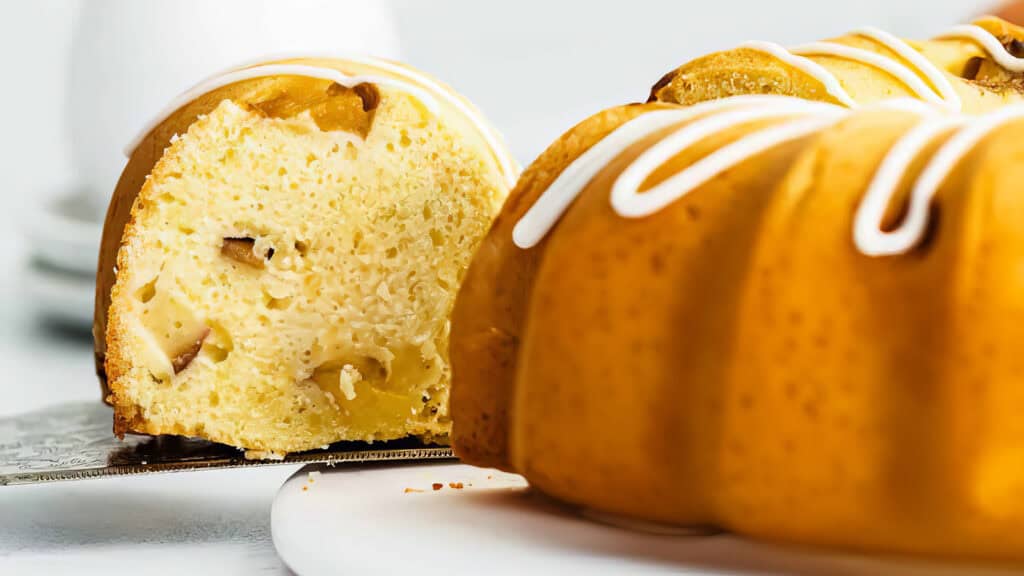 A close-up of a Bundt cake with a slice being lifted out. The cake is light yellow with visible pieces of peach through it.