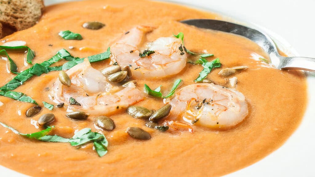 A creamy orange bisque infused with saffron, garnished with succulent shrimp, pumpkin seeds, and finely chopped basil. A silver spoon rests on the edge of the bowl, and a piece of bread is partially visible in the background.