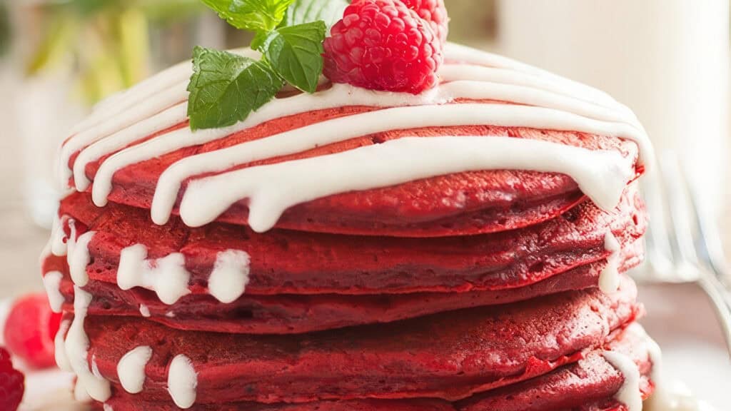 A stack of vibrant red velvet pancakes, topped with white icing, a fresh raspberry, and a sprig of mint. The background is softly blurred, focusing attention on the richly colored, tempting pancakes—a perfect addition to your favorite pancake recipes.