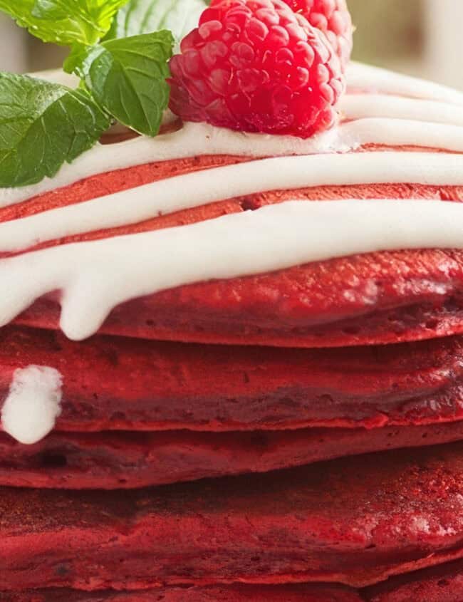 A stack of vibrant red velvet pancakes, topped with white icing, a fresh raspberry, and a sprig of mint. The background is softly blurred, focusing attention on the richly colored, tempting pancakes—a perfect addition to your favorite pancake recipes.