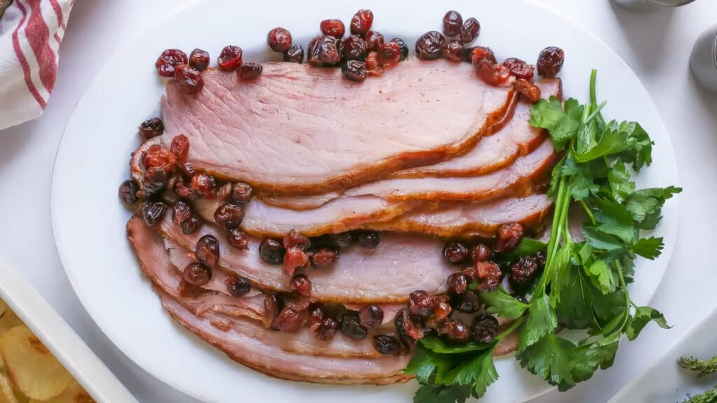 A platter of sliced ham garnished with cooked cranberries and sprigs of parsley, perfect for slow cooker recipes. The ham slices are arranged neatly on the white plate, surrounded by a few garnishes. The cranberries add a festive touch to this delightful dish.