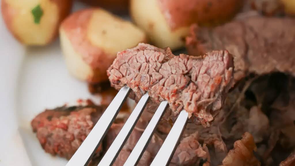 A close-up image of a fork holding a piece of cooked beef, a true highlight of slow cooker recipes. In the background, there are several small boiled potatoes with their skins on and more slices of beef. The focus is on the fork and beef in the foreground.