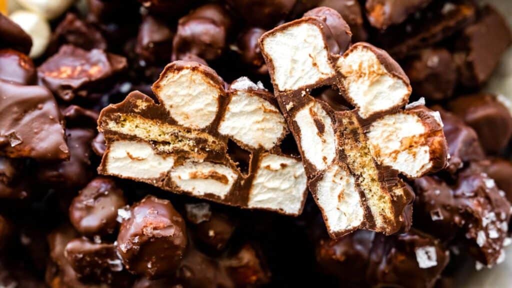 A close-up of a pile of rocky road candies, with one piece cut in half to reveal its sticky and sweet interior. The candy is made up of chocolate-coated marshmallows and nuts, giving a textured and mouth-watering appearance that's perfect for delicious recipes.