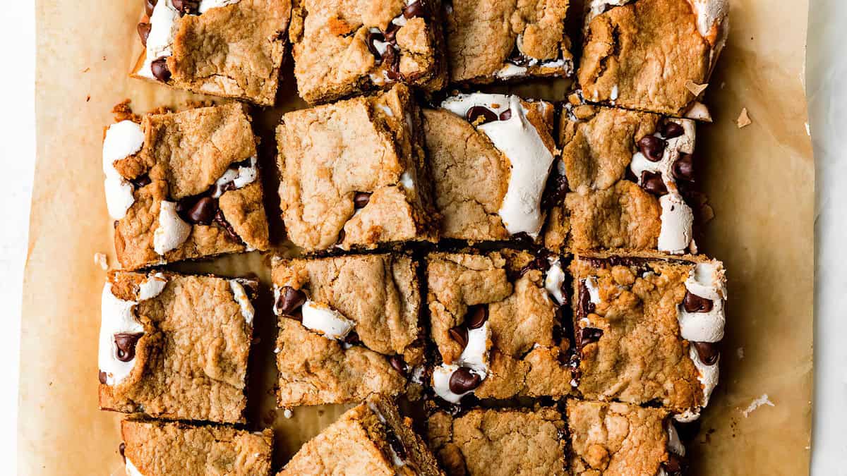 A close-up of 15 cookie bars arranged on parchment paper. The sweet treats have a golden-brown crust and are filled with chocolate chips and gooey marshmallow, creating an inviting, mouthwatering appearance.