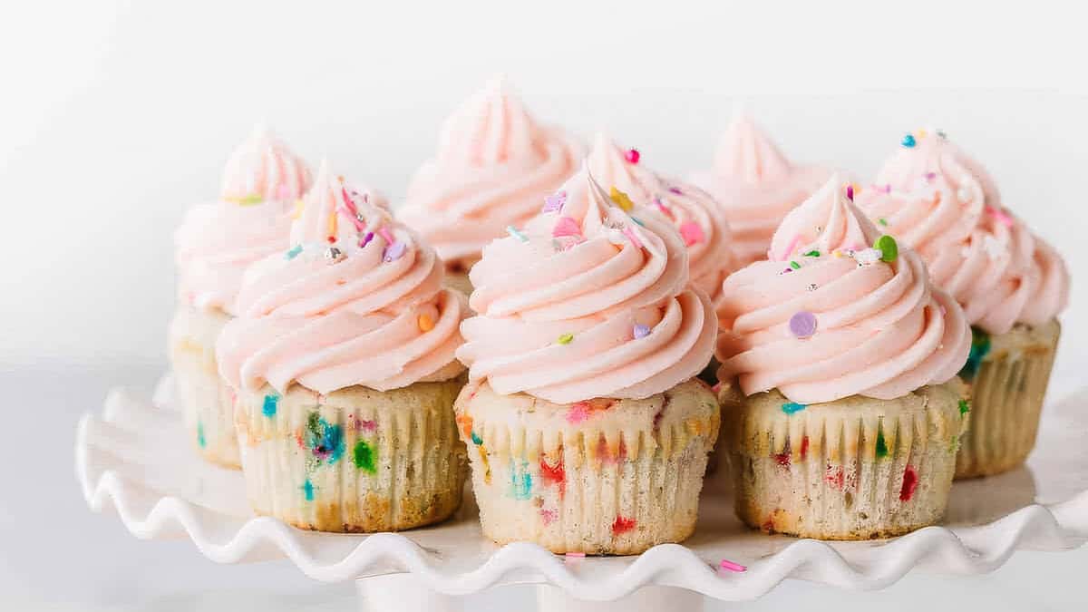 Seven cupcakes with pink, swirled frosting and colorful sprinkles are arranged on a white scalloped cake stand. The confetti cupcakes, set against a plain white background, delight the eye with their vibrant hues.