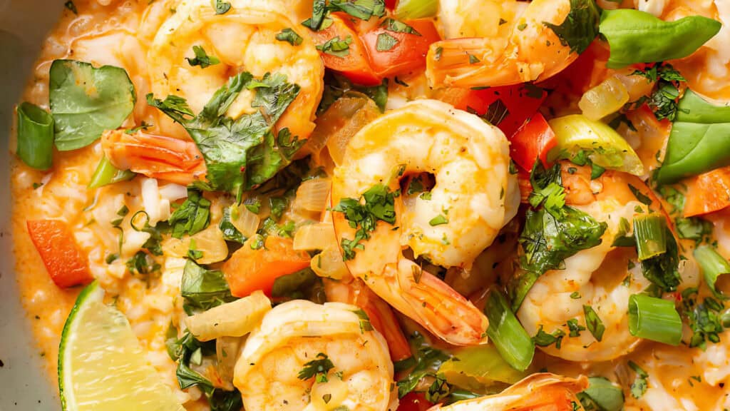 Close-up shot of a colorful shrimp dish featuring large, cooked shrimp, red bell peppers, green snap peas, and chopped fresh herbs. The meal is garnished with a lime wedge and served in a rich, creamy sauce—an enticing addition to your favorite Shrimp Recipes collection.