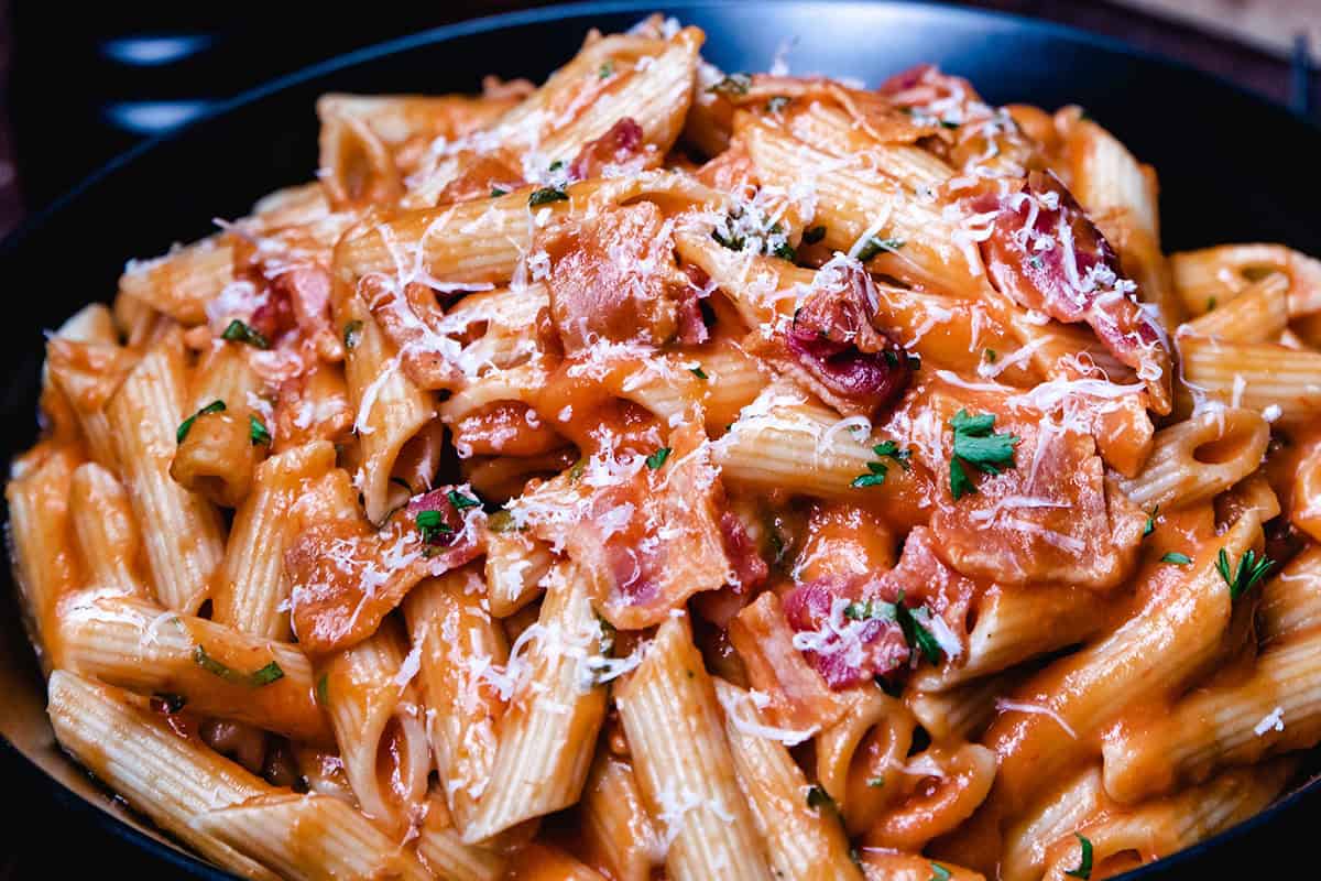 A close-up of a plate of creamy penne pasta mixed with chunks of crispy bacon, garnished with grated Parmesan cheese and chopped parsley. The rich, orange-colored sauce coats the pasta and bacon evenly, creating a delicious-looking dish that's perfect for easy dinner recipes.