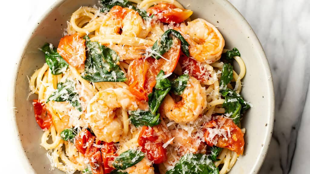 A bowl of spaghetti topped with succulent shrimp, sautéed spinach, and roasted tomatoes, garnished with grated Parmesan cheese. This vibrant dish is served in a white ceramic bowl against a white marble surface—perfect for those seeking delicious shrimp recipes.