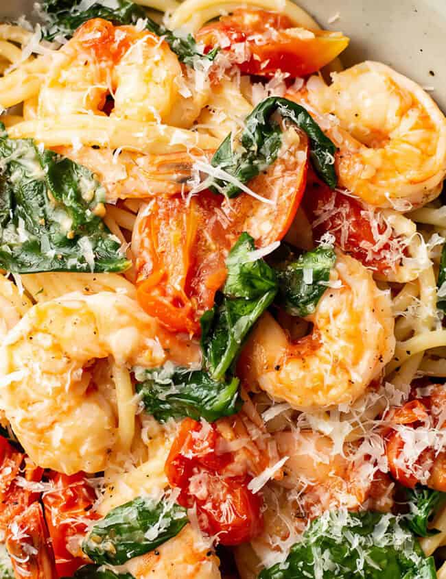 A bowl of spaghetti topped with succulent shrimp, sautéed spinach, and roasted tomatoes, garnished with grated Parmesan cheese. This vibrant dish is served in a white ceramic bowl against a white marble surface—perfect for those seeking delicious shrimp recipes.