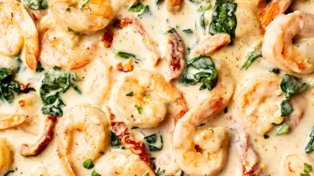 Close-up image of creamy Tuscan garlic shrimp. The dish features shrimp cooked in a rich, creamy sauce with spinach, sun-dried tomatoes, and garlic, garnished with fresh parsley. Perfect for those seeking delicious shrimp recipes, the sauce has a speckled appearance from black pepper and herbs.