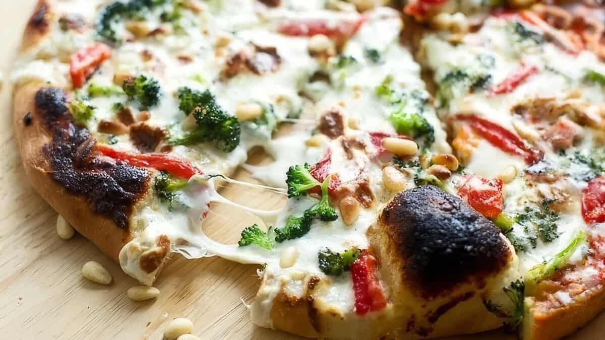 A close-up of a pizza slice being lifted from the whole pizza. Topped with melted cheese, broccoli, red bell peppers, and pine nuts, this golden brown crust is perfect for those who wish they could eat it every day. The background is a wooden surface.