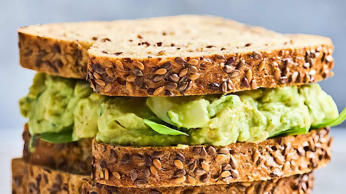 Close-up of a sandwich made with two slices of multigrain bread. The filling includes mashed avocado and fresh spinach leaves, with visible seeds on the bread crust. Perfect for breakfast or lunch, the background is slightly blurred, focusing attention on the delicious sandwich.