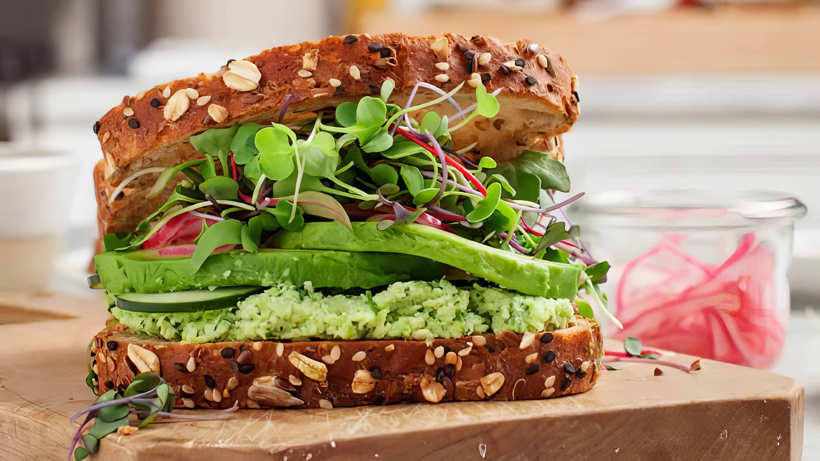 A hearty sandwich with whole grain bread, layered with fresh avocado slices, microgreens, cucumber, and a light spread on the bottom slice. The superfood avocado offers numerous health benefits. Garnished with additional microgreens, the sandwich is paired perfectly with a jar of pickled vegetables in the background.