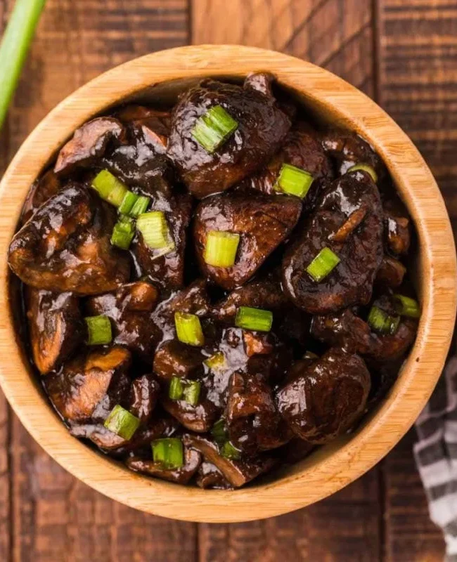 A wooden bowl filled with glazed mushrooms garnished with chopped green onions, set on a wooden surface. A bunch of green onions lies to the left, and a striped cloth napkin is partially visible on the right, perfect for those who love Asian recipes.