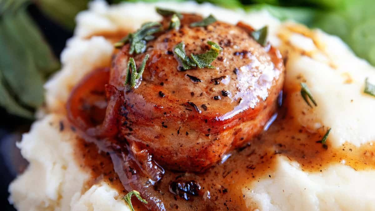 A close-up of a pork medallion wrapped in bacon, garnished with herbs, and topped with a savory brown sauce. This comfort food recipe features the medallion resting on a bed of creamy mashed potatoes with some greens visible in the background.