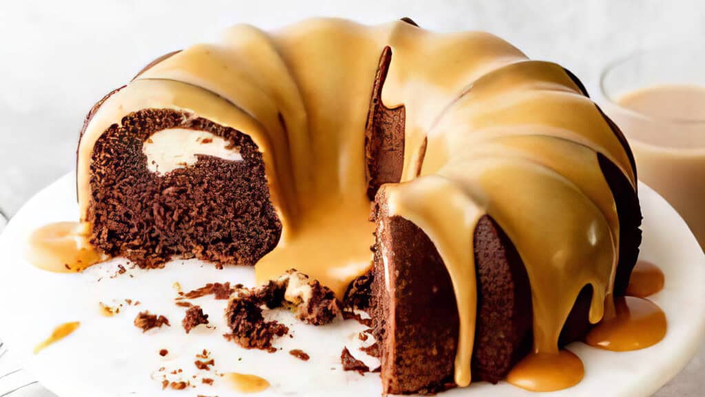 A chocolate bundt cake with a caramel glaze, partially sliced to reveal a creamy filling inside. Crumbs and cake pieces are scattered on a white surface around the cake, evoking classic Bundt Cake Recipes. A glass of a light-colored beverage sits blurred in the background.