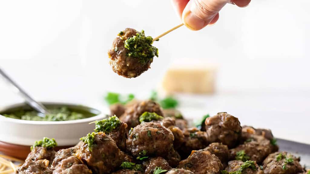 A hand holds a toothpick with a meatball covered in green herb sauce over a plate of more meatballs, showcasing one of the most delicious ground beef recipes. A bowl of additional herb sauce is to the left, garnished with fresh herbs. The background is out of focus, highlighting the food in the foreground.