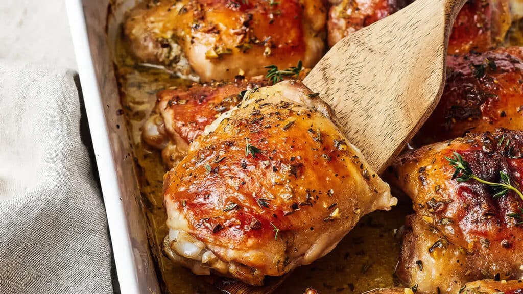 A close-up of golden-brown roasted chicken thighs in a baking dish, garnished with fresh thyme. A wooden spatula is lifting one piece, revealing juicy, tender meat and a perfectly crispy skin. A beige cloth napkin is partially visible on the side—an enticing addition to your favorite chicken recipes.