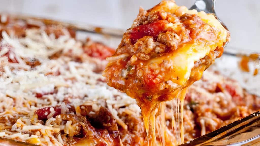 A close-up shot of a slice of lasagna being lifted from a glass baking dish, perfect for pasta lovers. The lasagna features layers of melted cheese, meat sauce, tomatoes, and pasta, with cheese stretching from the slice in the spoon and more grated cheese visible on the surface.