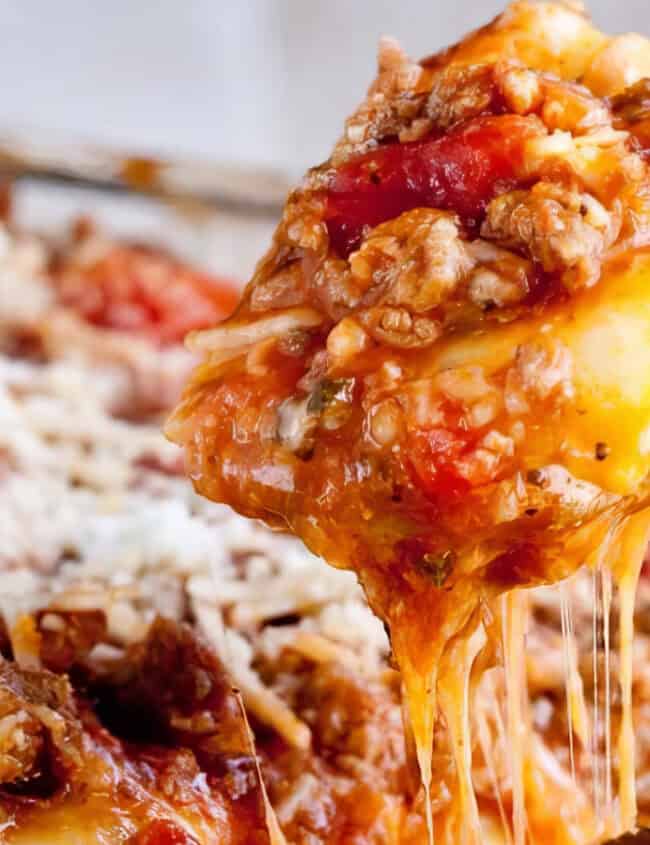 A close-up shot of a slice of lasagna being lifted from a glass baking dish, perfect for pasta lovers. The lasagna features layers of melted cheese, meat sauce, tomatoes, and pasta, with cheese stretching from the slice in the spoon and more grated cheese visible on the surface.