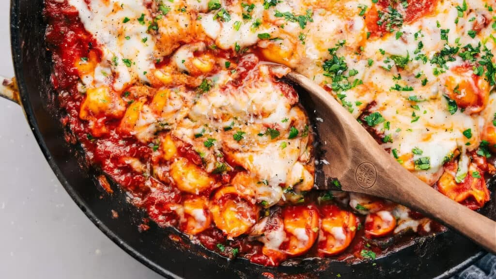 A close-up of a skillet filled with cheesy baked tortellini in a rich tomato sauce, garnished with chopped parsley. A wooden spoon is scooping some of the gooey, melted cheese and pasta, showcasing the dish's hearty and comforting texture—a delight for pasta lovers everywhere.