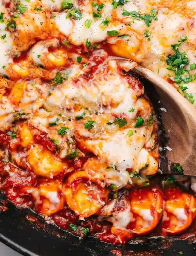 A close-up of a skillet filled with cheesy baked tortellini in a rich tomato sauce, garnished with chopped parsley. A wooden spoon is scooping some of the gooey, melted cheese and pasta, showcasing the dish's hearty and comforting texture—a delight for pasta lovers everywhere.