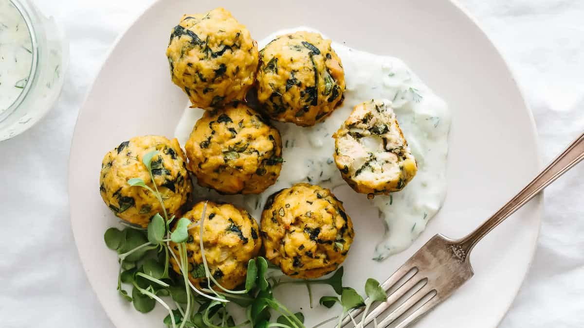 Six spinach and cheese balls on a white plate with a fork. The balls are accompanied by a creamy white dipping sauce and garnished with fresh greens. One ball is cut in half, revealing its interior—a true Meatball Magic moment!