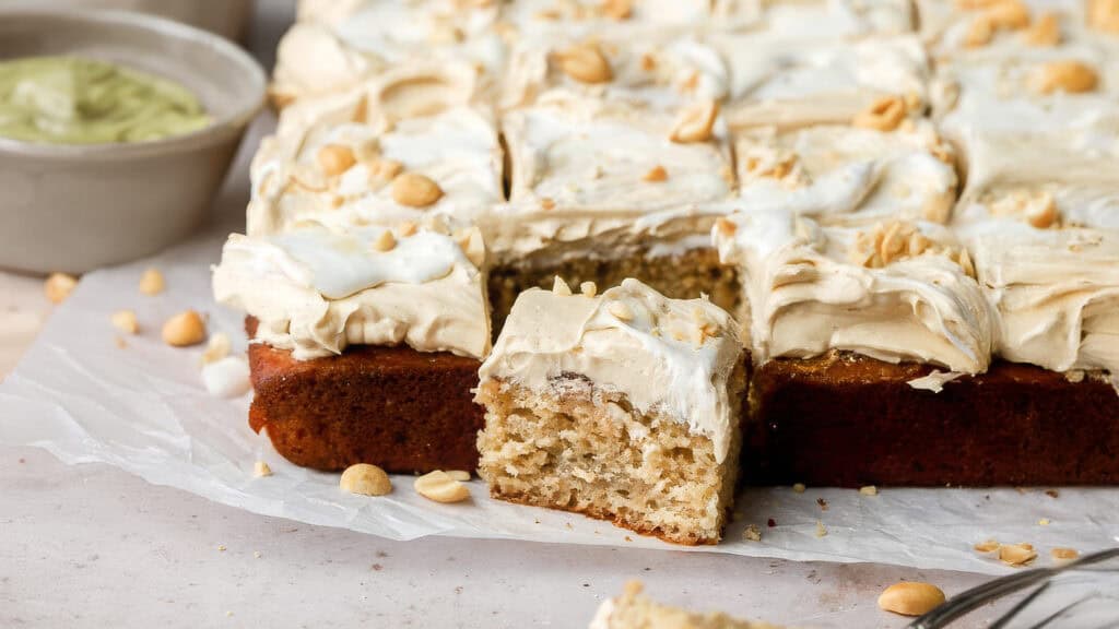 A sliced sheet cake with beige frosting, topped with chopped nuts, is presented on a piece of parchment paper. One piece is slightly pulled out, revealing a soft, moist interior. A small bowl with a green spread is visible in the background—perfect for those looking for irresistible sheet cake recipes.