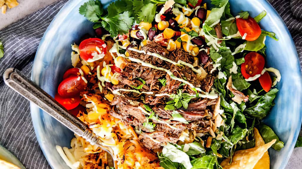 A vibrant bowl filled with shredded meat from one of our favorite slow cooker recipes, cherry tomatoes, corn, black beans, leafy greens, and a drizzle of creamy dressing, garnished with cilantro. There is a fork resting on the rim of the blue bowl.