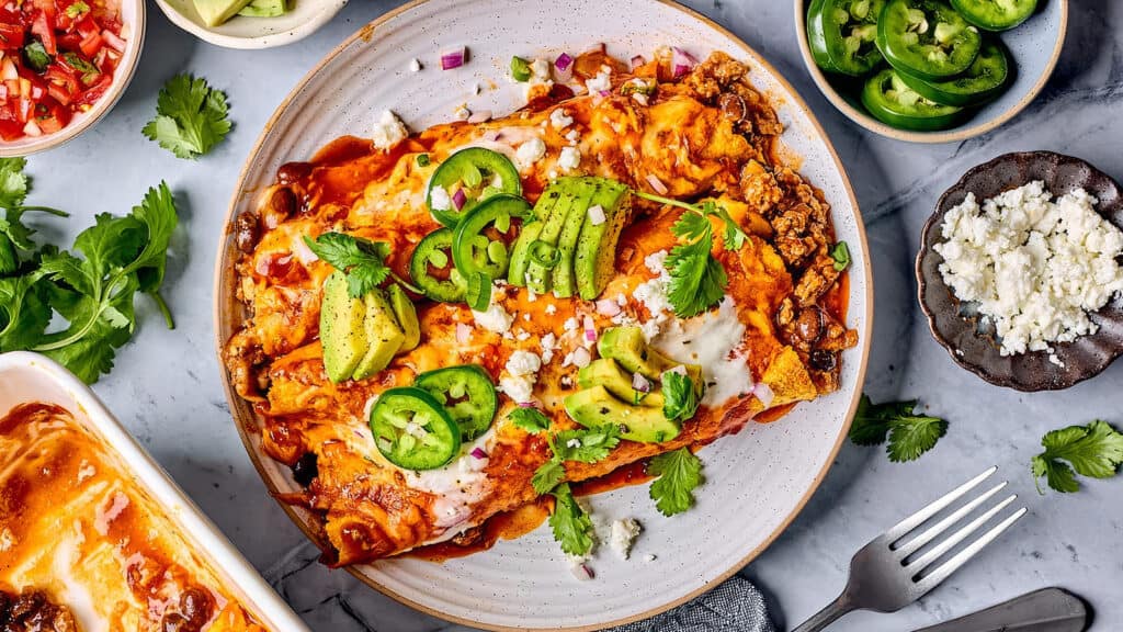 A plate of enchiladas topped with avocado slices, jalapeño slices, crumbled cheese, and fresh cilantro makes for a delightful centerpiece. Surrounding the main dish are bowls of various toppings, including diced tomatoes, sliced jalapeños, and crumbled cheese—perfect for exploring new ground beef recipes on a marble surface with a fork.