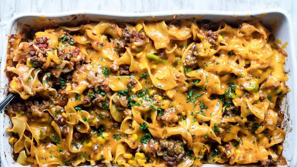 A baked casserole dish filled with cheesy ground beef and egg noodles, topped with melted cheddar cheese and sprinkled with chopped parsley sits enticingly on a white countertop—a perfect addition to your collection of ground beef recipes.