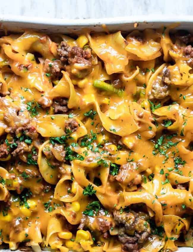 A baked casserole dish filled with cheesy ground beef and egg noodles, topped with melted cheddar cheese and sprinkled with chopped parsley sits enticingly on a white countertop—a perfect addition to your collection of ground beef recipes.