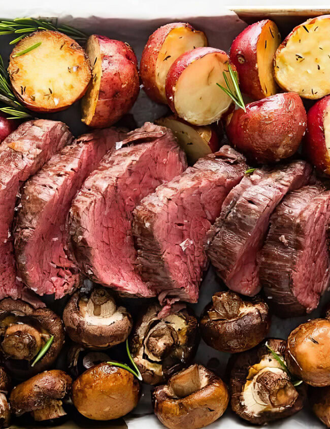 A tray with evenly sliced medium-rare roast beef in the center, surrounded by halves of roasted red potatoes garnished with rosemary, and a variety of roasted mushrooms. This delicious spread is an excellent addition to your sheet pan recipes collection. The tray is lined with parchment paper.