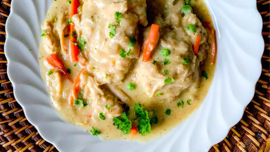 A white plate filled with creamy chicken and dumplings, featuring shredded pieces of chicken, carrots, and peas. This Southern recipe is garnished with fresh parsley and served in a rich, thick sauce, all placed atop a woven placemat.