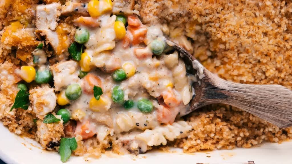 A close-up of a creamy chicken and vegetable casserole with a breadcrumb topping. The wooden spoon reveals mixed vegetables including peas, carrots, and corn, all enveloped in a rich sauce, with pieces of chicken visible throughout the dish—an ideal addition to your favorite casserole recipes.