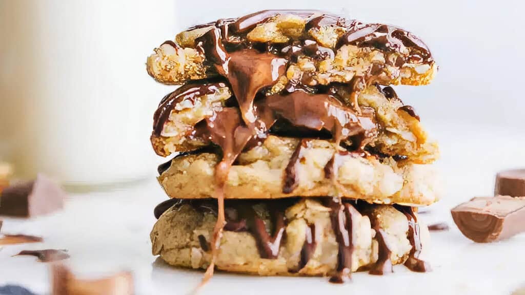 A close-up of three stacked chocolate chip cookies, drizzled with melting chocolate sauce. The cookies have chunks of chocolate and a gooey texture, reminiscent of the best caramel recipes. Blurred objects including more chocolate pieces and a glass of milk are visible in the background.