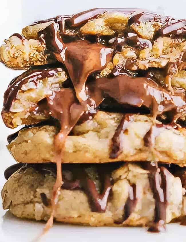 A close-up of three stacked chocolate chip cookies, drizzled with melting chocolate sauce. The cookies have chunks of chocolate and a gooey texture, reminiscent of the best caramel recipes. Blurred objects including more chocolate pieces and a glass of milk are visible in the background.