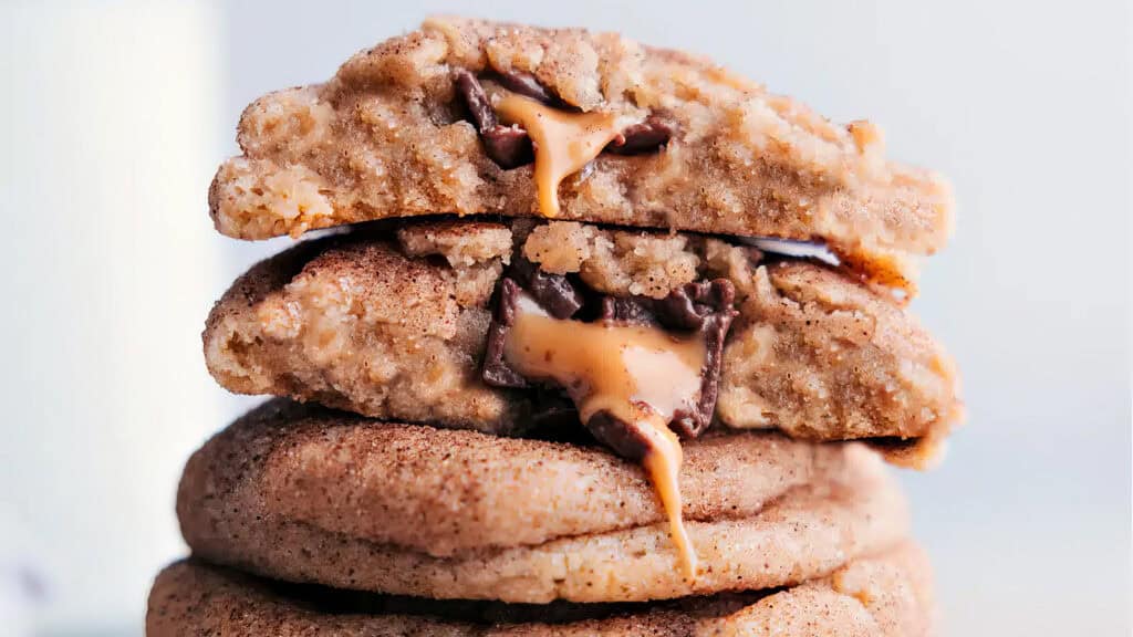 Stack of gooey chocolate and caramel-filled cookies, perfect for any caramel recipes. The cookies on top are split open, revealing melting chocolate and caramel inside. The outer layer of each cookie is light brown with a slightly crinkled texture. The background is blurred and neutral.