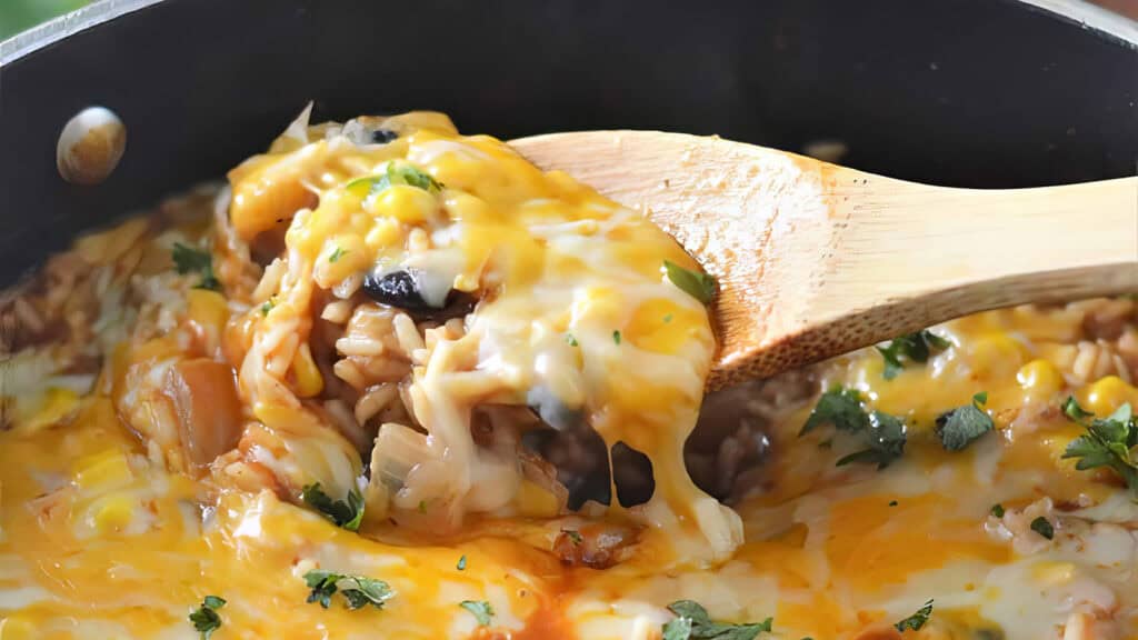 A close-up of a wooden spoon scooping a cheesy rice dish from a pan. The dish includes melted cheese, corn, black olives, rice, and is garnished with chopped cilantro. The mixture looks creamy and gooey with a savory, comforting appeal perfect for rice dish recipes.