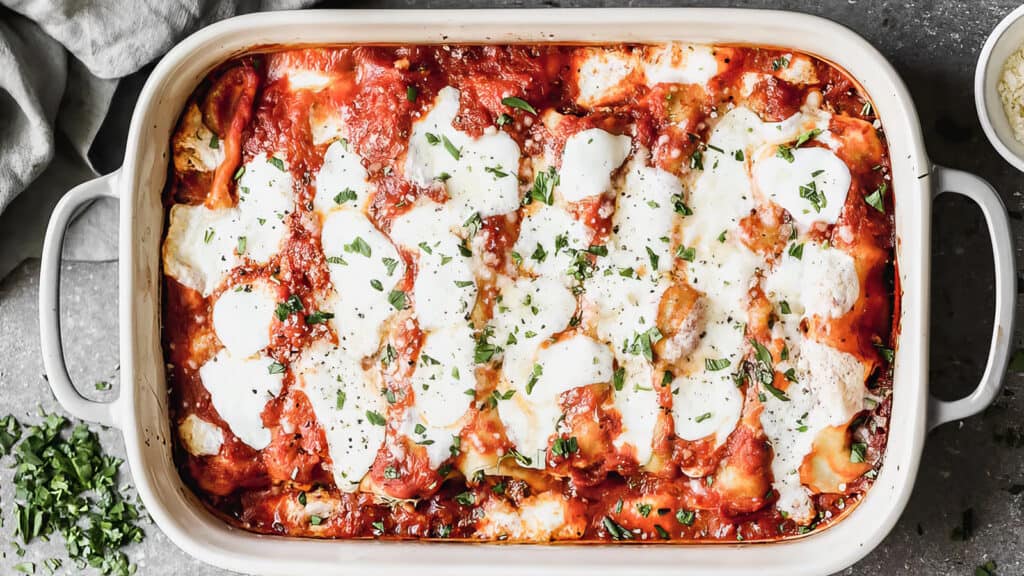 A rectangular baking dish filled with freshly baked lasagna, a delight for pasta lovers. The lasagna features layers of melted cheese, tomato sauce, and scattered herb garnish on top. The dish rests on a gray countertop, accompanied by small bowls of chopped herbs and grated cheese.