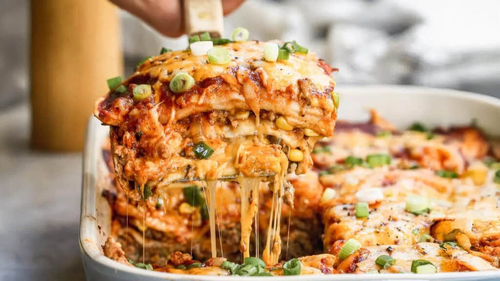 A close-up image of a cheesy casserole being served from a baking dish. The delectable casserole has multiple layers, including cheese, sauce, and visible green onions sprinkled on top. The cheese is melted and stretching as a piece is lifted with a spatula, making it an ideal candidate for your favorite casserole recipes.