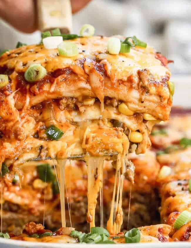 A close-up image of a cheesy casserole being served from a baking dish. The delectable casserole has multiple layers, including cheese, sauce, and visible green onions sprinkled on top. The cheese is melted and stretching as a piece is lifted with a spatula, making it an ideal candidate for your favorite casserole recipes.