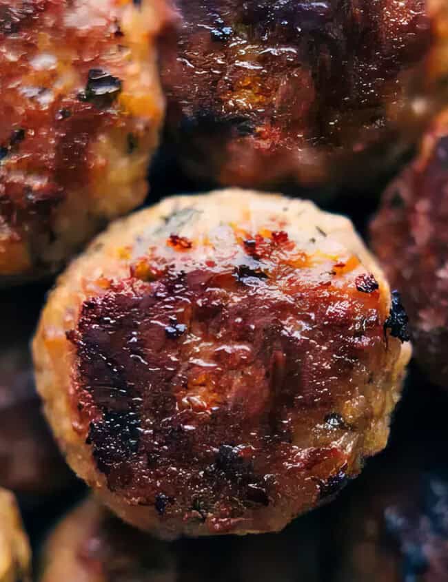 Close-up of a pile of cooked meatballs, featuring a crispy, golden-brown exterior. The meatballs show seared marks, suggesting they were grilled or fried. Some greens are visible in the upper left corner, hinting at a garnish or salad accompaniment. Discover more in our "Meatball Magic" guide.