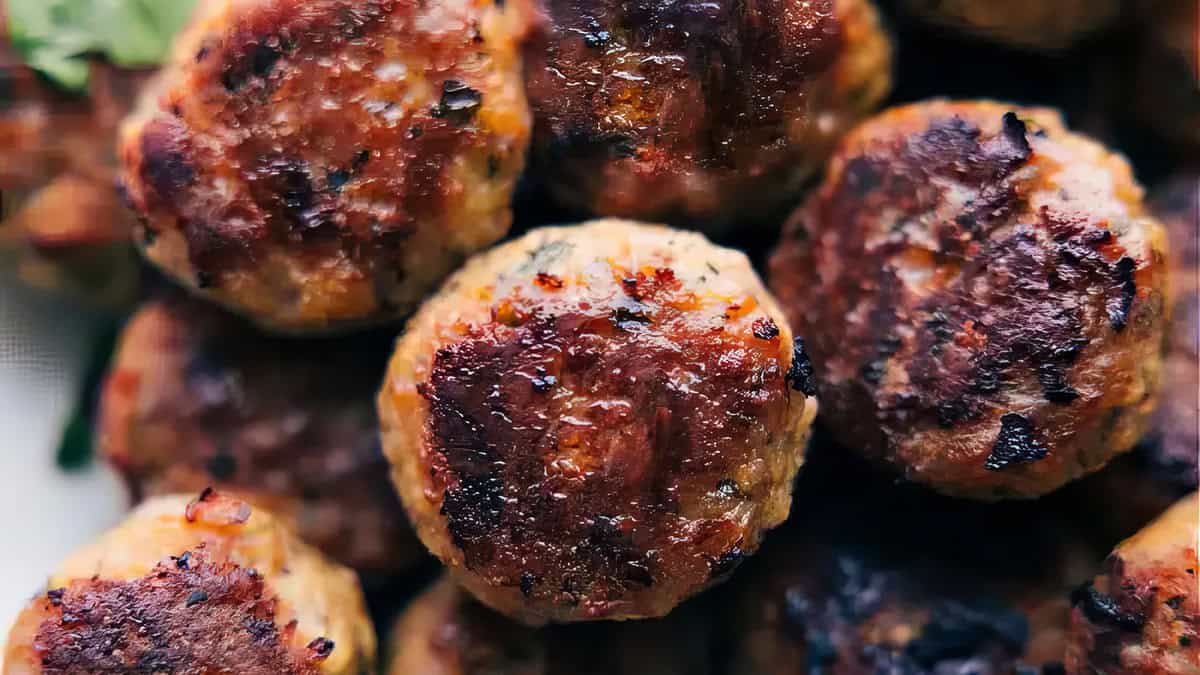 Close-up of a pile of cooked meatballs, featuring a crispy, golden-brown exterior. The meatballs show seared marks, suggesting they were grilled or fried. Some greens are visible in the upper left corner, hinting at a garnish or salad accompaniment. Discover more in our "Meatball Magic" guide.