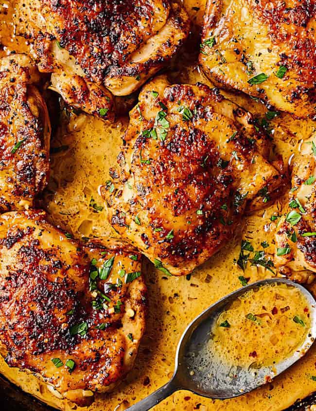 A skillet filled with cooked, golden-brown chicken thighs garnished with fresh herbs and a sprinkle of spices. A serving spoon rests in the spiced sauce surrounding the chicken. Fresh parsley is placed beside the skillet for garnish—perfect for those seeking delicious chicken recipes.